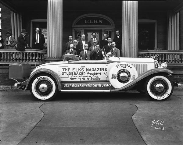 Elks Magazine purple and white free wheeling Studebaker President 8 roadster convertible parked in front of the Elks Club, 120-124 Monona Avenue. The car's crew is posing with local men who welcomed them. The automobile has a sign that reads: "The Elks Magazine - Studebaker President 8 - free wheeling from New York to Seattle."