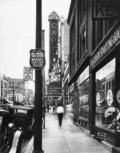 Pedestrians (in motion) are walking down State Street towards the Capitol building, near the Capitol Theatre marquee, RKO sign, the YWCA, Oettings Restaurant and Wisconsin Sporting Goods store. Text with print: "Looking east along 200 block of State St."