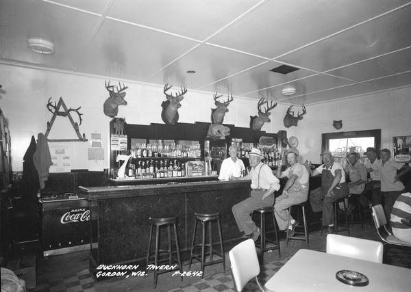 Customers sitting at the bar in the Buckhorn Tavern. There are five mounted deerheads above the bar. The bartender is standing behind the bar.