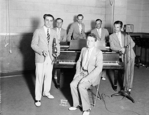 Anthony "Tony" Salerno's Gypsy Melodians at WIBA radio station standing around a piano. Tony is holding his violin, the other Melodians are standing around the piano, and Otto Heinz is sitting in front. Ben Ehr, sax and clarinet; Earl Barnes, trombone; Erwin Krueger, drums; and William Kraft, cornet.