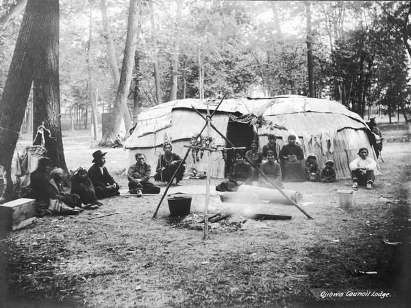 Ojibwa chief's lodge from Lac Courte Orielles Reservation set up at the 1906 Wisconsin State Fair. Note both reed and bark coverings on the lodge. A number of Indians — men, women and several children — sit in front of the lodge, and there is a fire pit in the foreground. Text at bottom right reads: "Ojibwa Council Lodge."