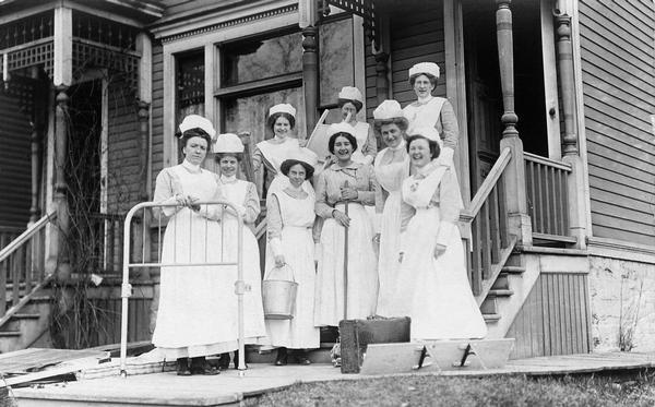 Eight nurses in uniform posing in front of a building (probably their workplace). Some of the women are holding a bedframe, pail, and a broom, and other items are displayed on the ground in front of the group.