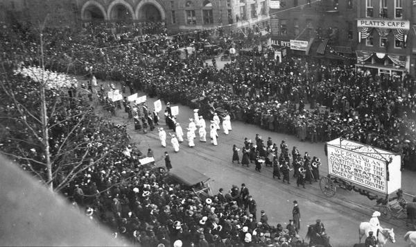 Elevated view of a suffrage parade, with a large crowd looking on. The sign on the horse-drawn wagon reads: "We Demand an Amendment to the Constitution of the United States Enfranchising the Women of this Country." A sign on the building across the street reads: "Platt's Cafe."