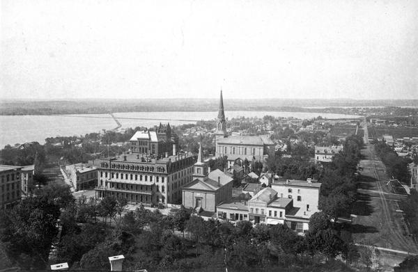 View from the Wisconsin State Capitol looking south. Lake Monona is to the left, and West Washington Avenue is on the right. The Dane County Courthouse on South Main Street is under construction, a fact which dates this picture. Also prominent is the tall spire of St. Raphael Catholic Church.  In the foreground are the Capitol Park and Carroll Street.  Facing Carroll Street are the Park Hotel and the First Baptist Church.