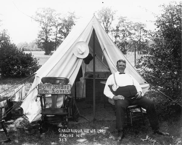 A man in bow tie and suspenders sits in front of a small tent. Next to him is a rocking chair with a sign on it that says "Don't you see I'm Lonely." Written onto the negative is "Chautauqua Views. 1906 Racine Wis, 363."
