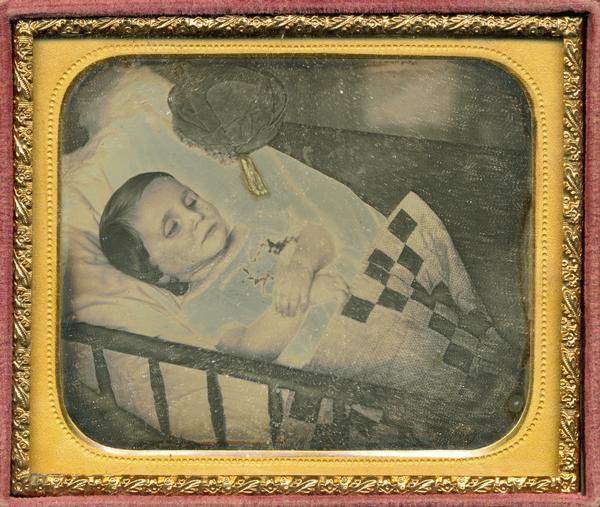 Sixth plate daguerreotype of a portrait of George Henry Williams (deceased child at 18 months), son of Henry C. and Diana T. Williams, and may be the cousin of N.M. Brown. The portrait shows the child laying in a cradle with a blanket and a small hat in the corner. Portions of the portrait have been tinted blue. The photograph was donated by Irving Brown in 1956.
