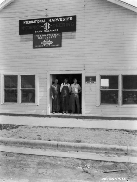 View from street towards three men standing in the doorway of an International Harvester farm equipment and truck dealership.