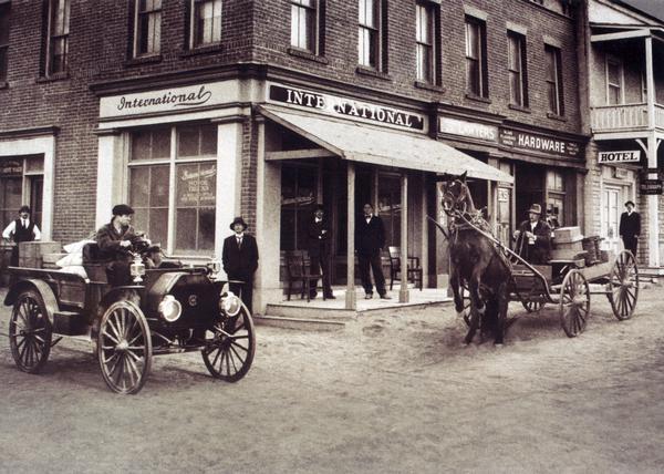Street corner featuring an International truck dealership, hardware store and hotel. Men are standing on the sidewalk in front of the dealership building. An International autowagon and a horse-drawn wagon are in the street. The horse appears to be rearing up at the sight of the autowagon.