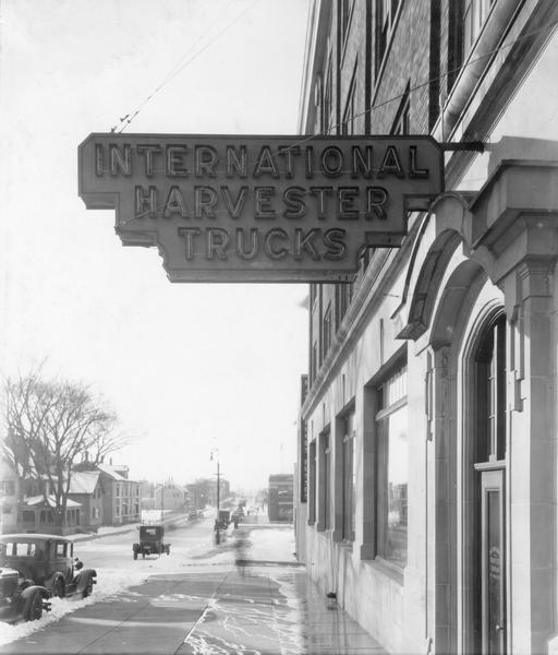 Cars and houses along a snowy street as seen from under a neon "International Harvester Trucks" sign.