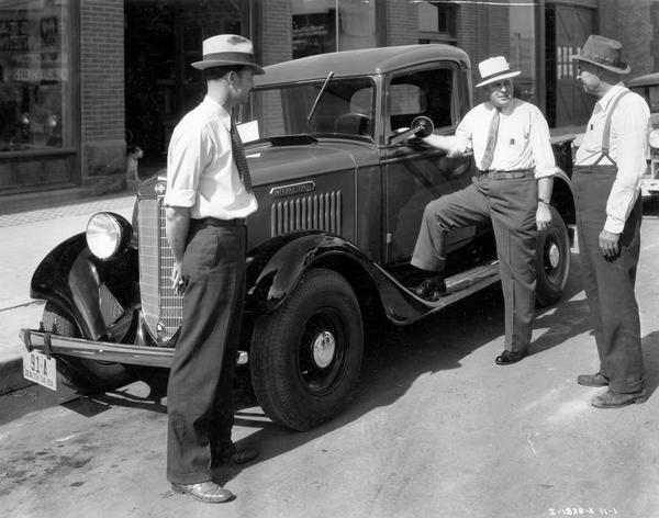 Three men standing around and International truck parked outside the dealership building of Ora McVey and Sons. The dealership sold International motor trucks and McCormick-Deering farm machines.
