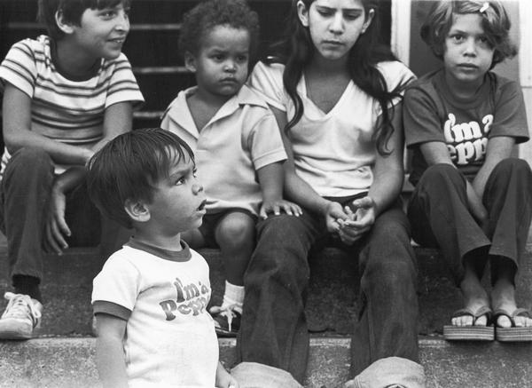 Children sitting on steps. Two of them are wearing "I'm A Pepper" t-shirts. Photographs made on July 4, 1980, by Archibald of Cuban refugees who had arrived as a result of the Port of Mariel exodus, and were housed at Camp McCoy, Wis.; including images of the camp life of Cuban men, women, and children.
