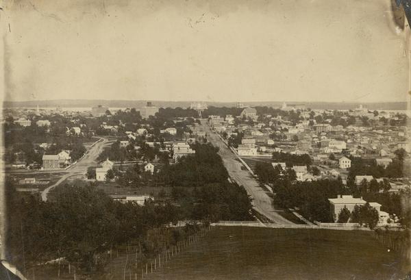 View of Madison from Main Hall, now Bascom Hall, on the University of Wisconsin-Madison campus looking up State Street toward the second Wisconsin State Capitol building. A portion of the lawn in front of Main Hall is in the foreground. Numerous residences dot the landscape, with several churches, Lake Mendota, Lake Monona, and the Farwell House in the distance.