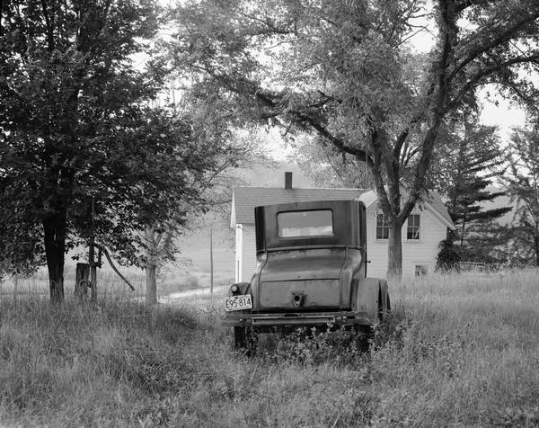 An old Ford automobile sitting in a farmyard, as viewed from the rear. There is a farmhouse in the background.