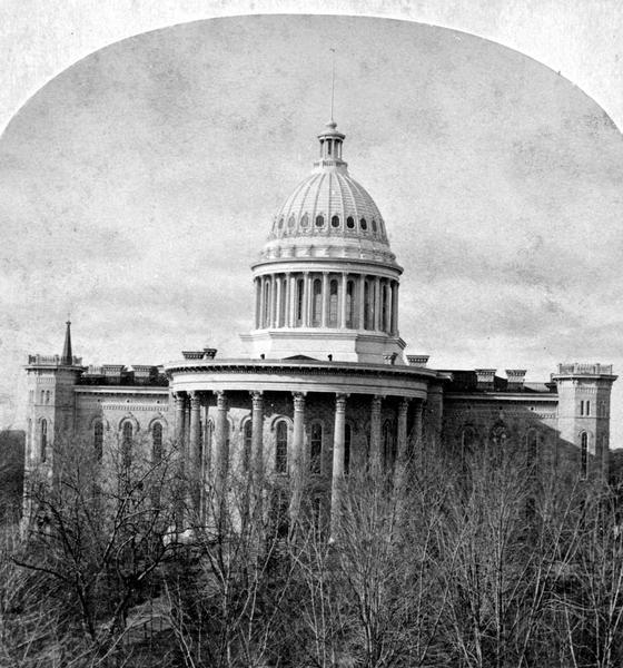Elevated view over trees toward the third (second in Madison), Wisconsin State Capitol that stood from 1857-1913, from a stereograph original. Elevated view over bare trees.