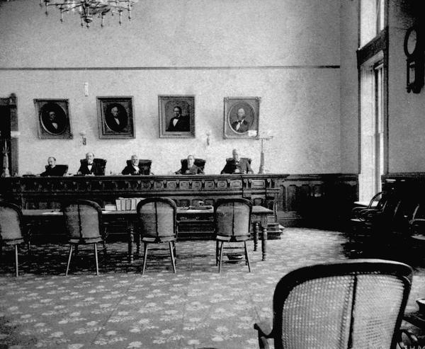 Interior view of the Supreme Court room in the third Wisconsin State Capitol, with Chief Justice William Penn Lyon seated in the center, and Justices J.B. Winslow, John B. Cassoday, Harlow S. Orton, and Silas U. Pinney around him. This photograph shows the impressive two-story  courtroom in the North Wing that was occupied by the Supreme Court after the expansion of 1883.