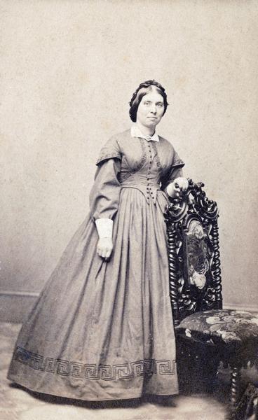 Formal studio photograph of a woman standing next to a chair.