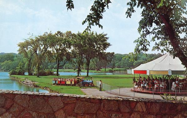 View from a stone bridge towards Vilas Park, with children riding the carousel (merry-go-round) and a kiddie train adjacent to the lagoon.