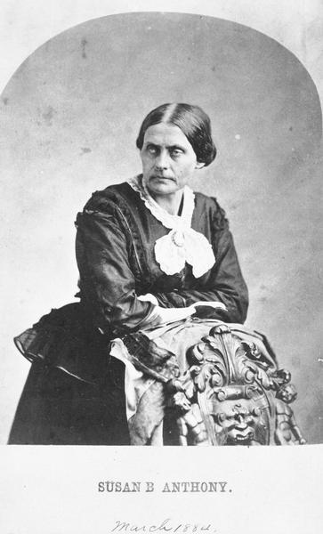 Studio portrait of Susan B. Anthony wearing a black dress. She is standing and leaning on the back of a chair.