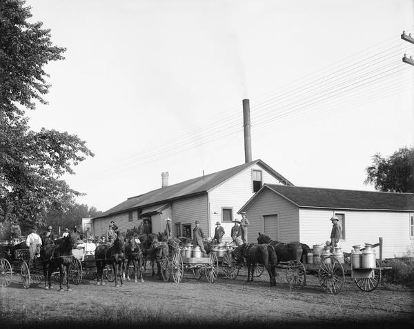 Farmers and horse-drawn wagons loaded with milk cans pose in front of a Wisconsin creamery.