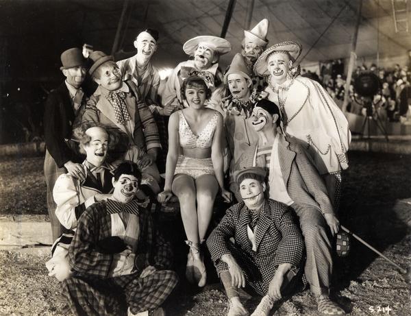 A group of 11 circus clowns poses with a pretty young female performer in a circus tent. This could possibly be part of a movie set.