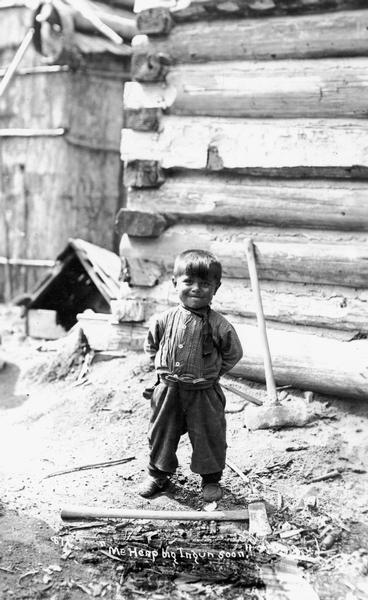 A Menominee child, probably photographed on the reservation near Keshena and Neopit, Wisconsin. He is standing outside in front of a log cabin, and an axe is on the ground in front of him. This image is part of an exhibit about Native Americans prepared by Paul Vanderbilt, the Wisconsin Historical Society's first curator of photography.