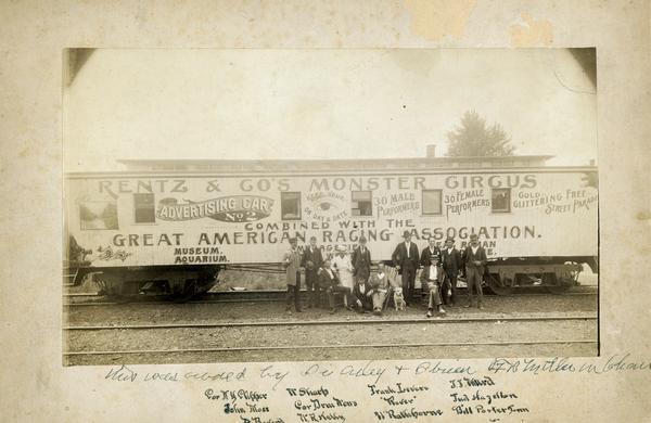 A group of personnel with the Rentz & Co.'s Monster Circus combined with the Great American Racing Association, pose for a photograph by the side of the train car advertising the show.