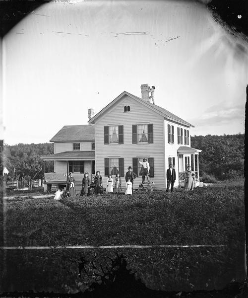 Harold and Gunhild Mickelson with five of their daughters (Bertha Mickelson Bragger and her husband are standing on the right), in Section 23 of the Town of Blue Mounds. One man is standing up on sawhorses in the center of the group holding a hand drill. A young child and a dog on the far left are blurred by movement. There is a man standing on the roof near the chimney.