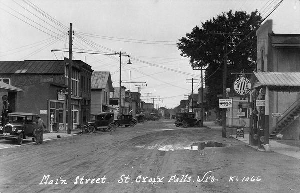 Main Street, with two competing curb-side gas stations across the street from each other. Cars are parked along both sides of the muddy street.  Such curb-side service stations were typical of the early days of automobiles.  Perhaps the large mud puddle in front of the Sinclair gas pumps has given the advantage this time to the station on the left.