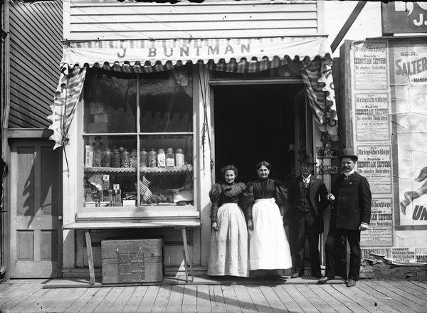 Two well-dressed women in aprons and two smartly dressed men pose in front of the open entrance of J. Buntman's fruit and confectionery store at 1005 North Eighth Street.  Jars of penny candy, tins of spices, American flags and boxes of fruit are some of the items displayed in the window.  Beneath a fold-down table, signs on a large wooden box advertise Old Soldier brand tobacco.  On the right, billboard posters advertise coverage of all the latest  news dispatches from home and abroad regarding the theater of war by the <i>Tagliche Sheboygan Zeitung</i>, the city's daily German newspaper.  "Theater of war" most likely refers to the Spanish-American War.