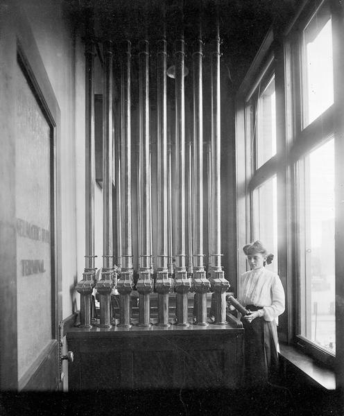 A worker stands next to a pneumatic tube communication system used at the Milwaukee Electric, Railway, and Light Company.