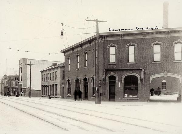 View across street towards the Hausmann Brewing Company on the corner of State and Gorham Streets. An archway on the right side leads to an inner courtyard, where a man is standing by a horse-drawn sleigh. It was razed in 1923-4. Handwriting at top of building reads: "Hausmann Brewing Co".
