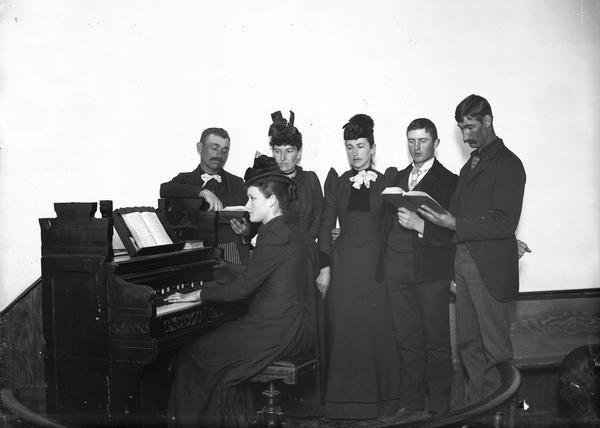 A woman plays the organ, while two women, wearing hats, and three men, gather behind her, holding song books. They are enclosed by a low, circular railing; the wall behind is blank.