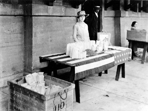 A member of the Dane County Council of Defense Food Board Women's Committee sells dried corn at the farmer's market. Women's organizations sold wheat substitutes to insure compliance with food conservation measures.  Over 3000 lbs. were sold in 1918. In 1917, the Catholic Women's Club installed a food drier at the market, which they operated as a patriot's work.