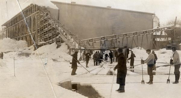 Men harvesting ice at the Conklin Ice House on Lake Mendota. The men are using pike poles to slide the blocks to the conveyor and U-shaped tools to split partially sawed blocks from ice. The steel frame of the new Wisconsin State Capitol is visible to the right of the icehouse.