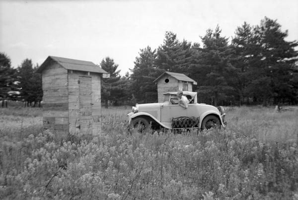 A man is driving an automobile in a field near two buildings, perhaps outhouses.
