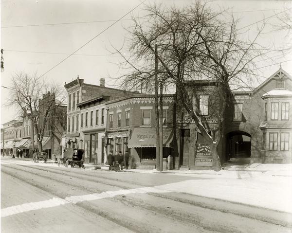View of T. Gullman Shoes, Simon Brothers Vegetables, and H.J. Minch Flour, Feed, Grain and Hay on the north corner of the 400 block of State Street. There is a blanket of snow on the ground, and men are walking under the awnings of the store fronts.