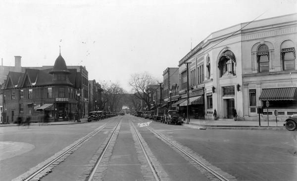 Intersection of Broom Street, Gilman Street and State Street with many storefronts like Lewis Prescription D, rugs, and marquees for Laundry, Delicatesen, McKillop's, Harnoff Loprich Electric Co. Also shows automobiles parked curbside on State Street, power poles and streetcar tracks. In the background is a view of Bascom Hill on the University of Wisconsin-Madison campus.