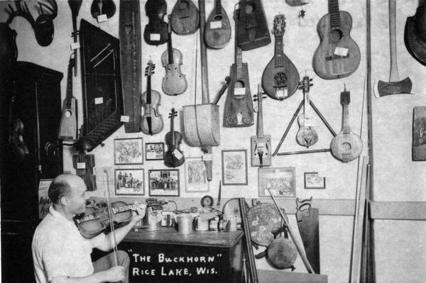 Otto Rindlisbacher, folk singer and maker of stringed instruments, and violin collection, sitting in his shop holding a Hardanger fiddle. Caption at bottom reads: "'The Buckhorn' Rice Lake."