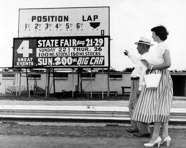 Mary Ellen McCabe, the Alice in Dairyland from Ladysmith, views a billboard announcing the stock car races scheduled for the Wisconsin State Fair during August 21-29. She is accompanied by an unidentified man.