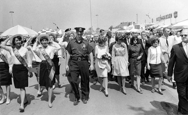 Beth Bartosh of Burlington, the 1964 Alice in Dairyland, walks along  the grounds of the Wisconsin State Fair, accompanied by a large group of people, including Lucy Baines Johnson, the daughter of President Lyndon B. Johnson, several supporters of Senator William Proxmire, a couple of police officers, and others. Lucy Baines Johnson walks on Alice's left.