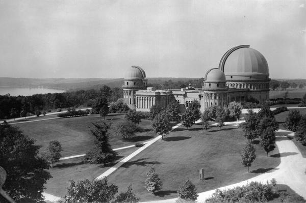 Elevated exterior view of Yerkes Observatory. Geneva Lake is in the background, left.  Roads and sidewalks cross the sweeping, well tended lawns.  The design of the grounds was based loosely on a plan by the firm of landscape architect Frederick Law Olmsted; the full plan was never implemented.  The grounds include several trees which are the largest of their species in Wisconsin and other interesting specimens.