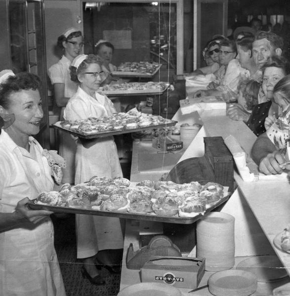 Four female bakers are holding up large trays laden with fresh cream puffs at a State Fair midway concession stand, while an eager crowd stands waiting in anticipation on the other side of the counter.