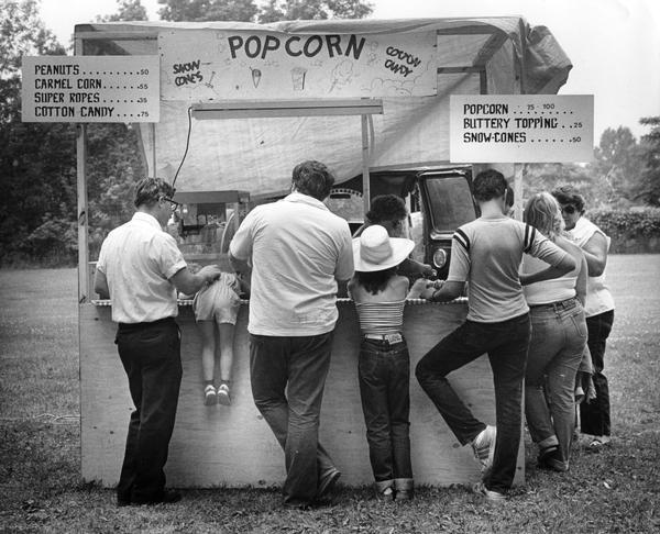 People, including a very eager little girl, stand around a concession stand to buy popcorn, peanuts, cotton candy, snow-cones, and other snacks at the 50th Annual Folk Fest/Volksfest, sponsored by the United German Societies of Milwaukee at Old Heidelberg Park.