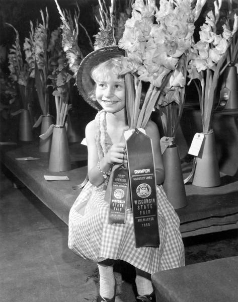 A smiling girl poses with stalks of gladiolas that won the champion ribbon for "Gladiolus Spike," and the first premium ribbon for "Horticulture Plants and Flowers" at the Wisconsin State Fair.