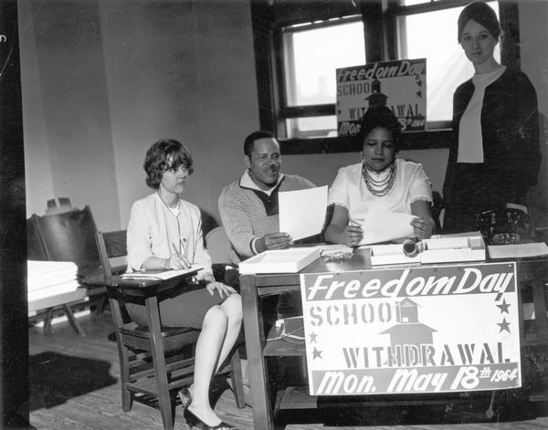 Four people seated at a desk which has a poster on it advertising Freedom Day School Withdrawal, set for Monday, May 18th, 1964.