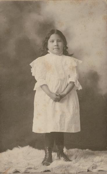 Studio portrait in front of a painted backgroud of Zintka Lanuni Colby as a little girl, wearing a white dress.