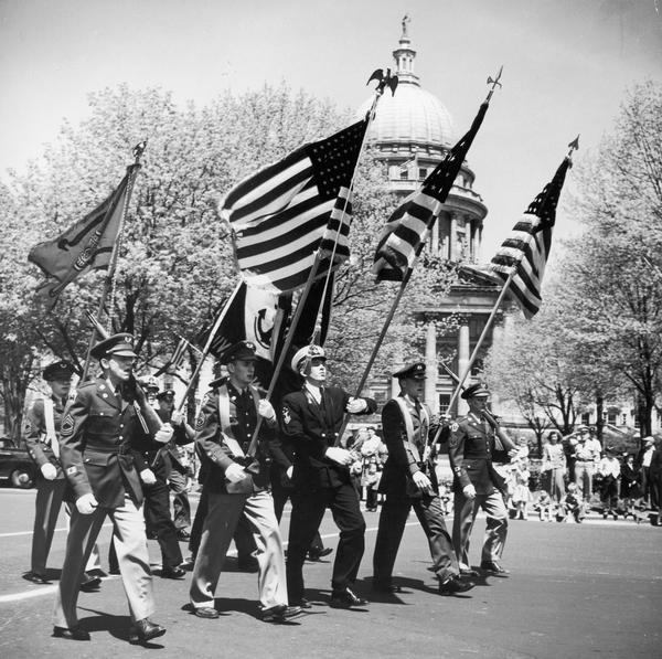 Color guards marching past the Wisconsin State Capitol carrying flags as a crowd looks on.