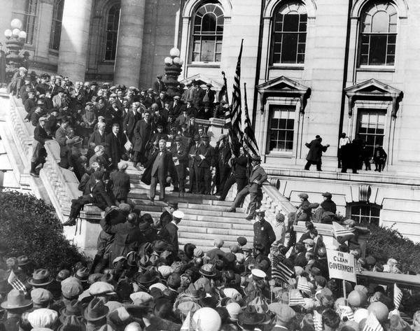 Robert M. La Follette, Sr. speaking to a large crowd from the Wisconsin State Capitol steps. This speech marked the end of his independent campaign for the presidency.  Photographers are perched on window sills and members of the crowd are waving flags and campaign signs.