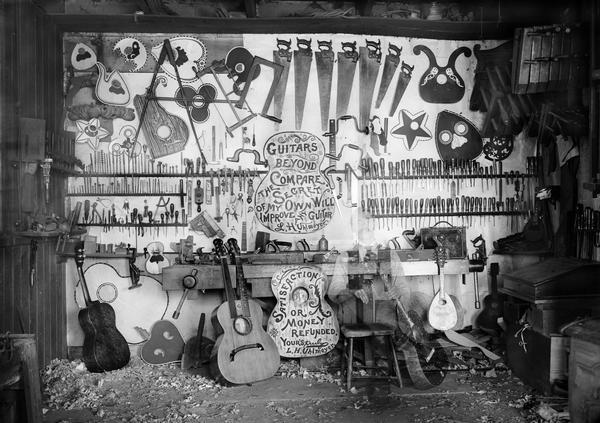 Interior of a guitar  maker's workshop, with a workbench, tools,  guitars, and other stringed instruments. In the foreground, there is a ghostly image of a seated man, likely that of the luthier, Leo Uhlmeyer. This effect was created by the photographer's use of a long exposure, and having Uhlmeyer leave the scene before the exposure was completed.