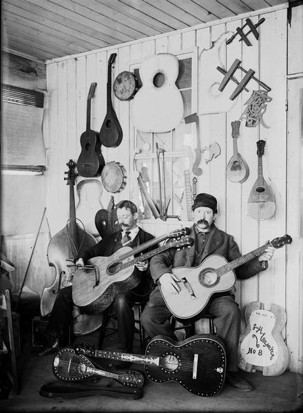Two men are seated holding guitars in luthier Leo Uhlmeyer's store. There are more instruments on the floor in front of them and hanging on the wall behind them.